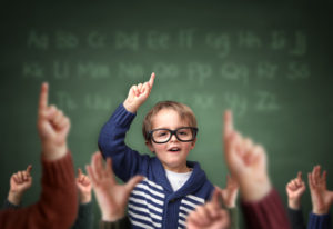 School child with hand raised in the classroom in front of a blackboard with other children concept for teacher's pet, standing out from the crowdand, genius or excelling in education